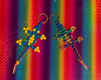 May include: Two beaded lizard keychains, one blue and yellow, the other multi-colored, on a rainbow background.