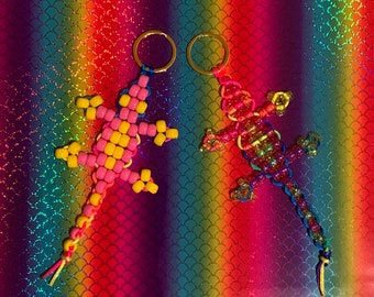 May include: Two beaded lizard keychains, one pink and yellow, the other with rainbow colored beads. The keychains are hanging on a rainbow background.