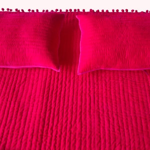 May include: A bright fuchsia bedspread with two matching square pillows. The bedspread has a textured, quilted design with vertical lines. The top edge is decorated with small, round pom-poms. The pillows have a similar quilted texture and a contrasting pink border.