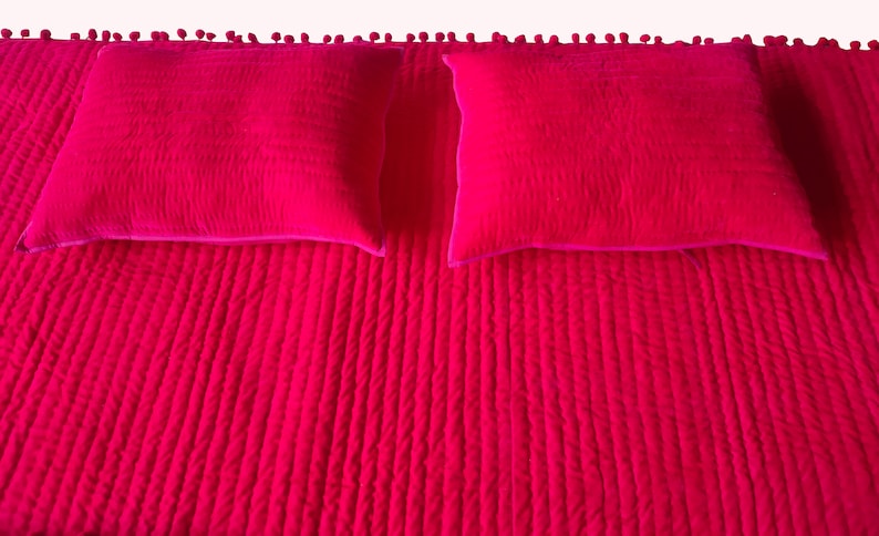 May include: A bright fuchsia bedspread with two matching square pillows. The bedspread has a textured, quilted design with vertical lines. The top edge is decorated with small, round pom-poms. The pillows have a similar quilted texture and a contrasting pink border.
