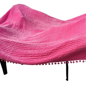 May include: A bright pink quilted blanket with a textured surface, draped over a dark-legged table. The blanket is trimmed with small pink pom-poms, adding a decorative touch to the furniture.