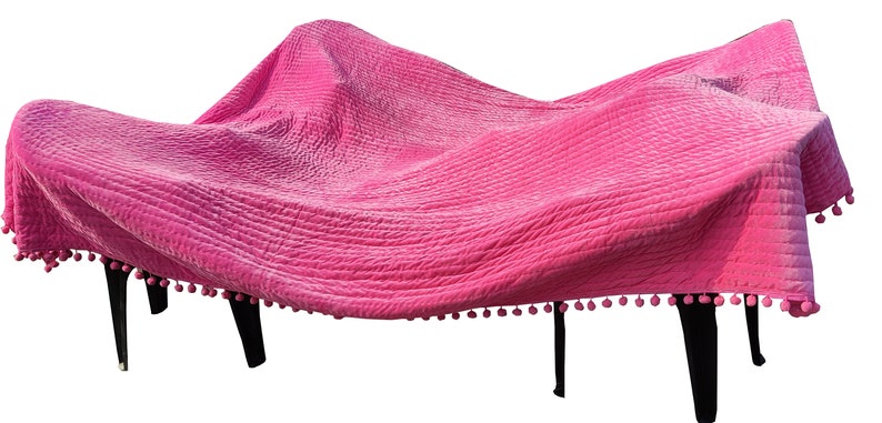 May include: A bright pink quilted blanket with a textured surface, draped over a dark-legged table. The blanket is trimmed with small pink pom-poms, adding a decorative touch to the furniture.