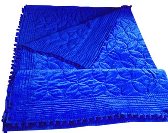 May include: A vibrant blue velvet quilt with a floral pattern and a pom-pom trim. The quilt is folded, showcasing the detailed stitching and plush texture. The bright color and design suggest a cozy home decor item.