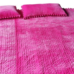 May include: A vibrant pink quilted bedspread with two matching pillows. The bedspread and pillows have a textured, crinkled look. The rectangular pillows are trimmed with small pink pom-poms. The bedspread is a solid colour with a subtle ombre effect.
