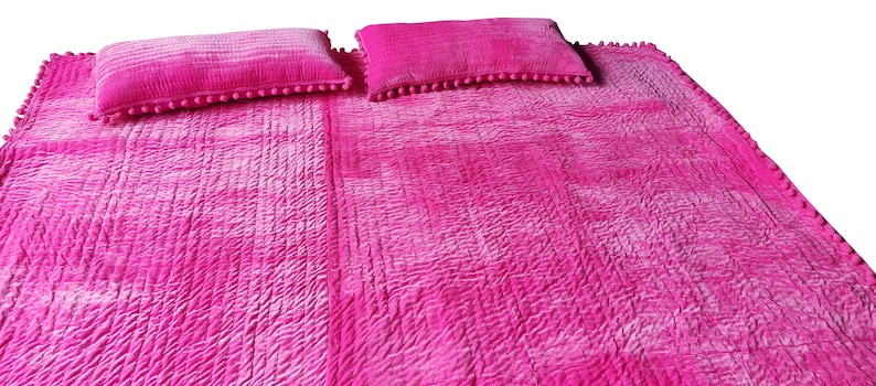 May include: A vibrant pink quilted bedspread with two matching pillows. The bedspread and pillows have a textured, crinkled look. The rectangular pillows are trimmed with small pink pom-poms. The bedspread is a solid colour with a subtle ombre effect.