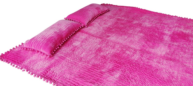 May include: A bright pink bedspread with two matching pillows. The bedspread and pillows have a textured, quilted appearance and are trimmed with small pink pom-poms. The set is designed for a bed or lounging area.