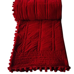 Puede incluir: Manta de tiro acolchada de terciopelo rojo con ribete de pompones. La manta est&aacute; doblada por la mitad y los pompones son visibles en ambos lados.