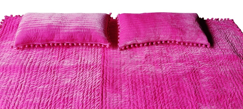 May include: A vibrant pink bedspread and two matching pillows. The bedspread and pillows feature a quilted design. The rectangular pillows are trimmed with small pink pom-poms. The bedding is a solid, bright colour.