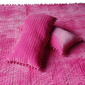 May include: A vibrant pink quilted throw and two matching rectangular cushions. The blanket and cushions feature a textured, stitched design and are trimmed with small pink pom-poms. The bedding is a solid colour.