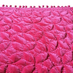 Puede incluir: Una manta de tiro acolchada de color rosa con un borde de pompones.