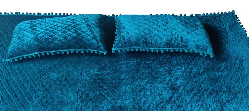 May include: A teal bedspread set with two matching pillows. The bedspread and pillows feature a quilted diamond pattern and are trimmed with small, matching pom-poms. The fabric has a crushed velvet texture.