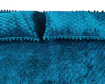 May include: Two teal blue velvet pillows with pom pom trim on a teal blue velvet bedspread.