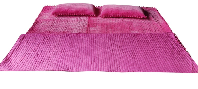 May include: A bright pink bedspread set with two matching pillows. The bedspread has a textured, quilted design and a solid pink border. The pillows are decorated with small pink pom-poms along the edges, adding a decorative touch to the bedding.