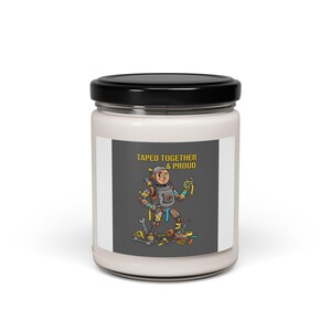 May include: A white candle jar with a black lid. The label features a cartoon astronaut with tools and the text "Taped Together & Proud". The astronaut is surrounded by tools, with a gray background.