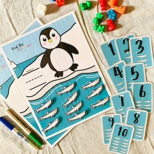 Op de afbeelding: Een printbaar educatief spel voor kinderen met een cartoonpinguïn op een ijsberg omringd door vissen. Het spel bevat nummerkaarten met visillustraties en een werkblad om nummers te schrijven.