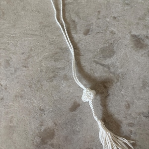 May include: A white tassel with a knotted detail and a loop at the top. The tassel is made of many thin, white cords. The background is a light gray, speckled surface.
