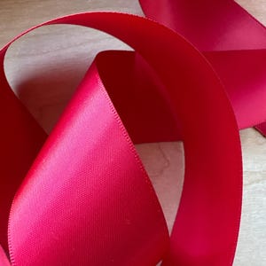 May include: Close-up of a vibrant red satin ribbon, its glossy surface reflecting light. The ribbon is curled and looped, showcasing its smooth texture and rich color. The ribbon's edges are neatly finished, suggesting a high-quality craft material.