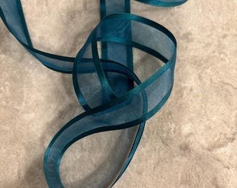May include: A teal sheer organza ribbon with solid satin edges. The ribbon is looped and twisted, showcasing its translucent fabric and deep blue-green colour. The material is lightweight and semi-transparent, creating overlapping layers of colour.