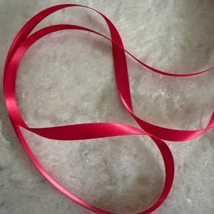May include: A single length of smooth, vibrant red satin ribbon arranged in a loose, flowing loop. The ribbon features a consistent, reflective finish on its surface, creating gentle curves and soft folds as it rests in a continuous, elegant shape.