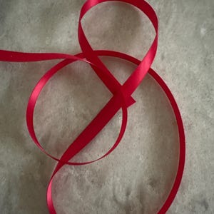 May include: A single length of smooth, vibrant red satin ribbon arranged in a loose, flowing loop that crosses over itself to create an open, organic shape with soft curves and a reflective, glossy finish along the edges of the fabric.