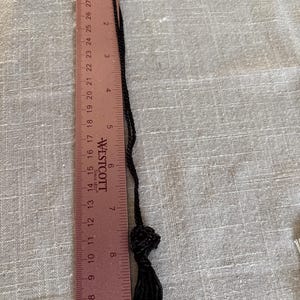 May include: A black tassel necklace with a knotted detail, measuring approximately 10 inches in length. The tassel is made of fine, black threads. A ruler is placed next to the necklace for measurement.
