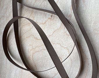 May include: A length of dark brown ribbon with a distinct ribbed texture. The ribbon is flexible and is shown in a loose, coiled arrangement, displaying its consistent width and matte finish throughout the entire length of the material.