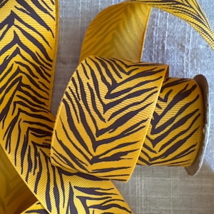 May include: A spool of yellow ribbon with a dark brown zebra stripe pattern. The ribbon is partially unrolled, revealing the pattern and texture. Ideal for crafts, gift wrapping, and decorative projects.