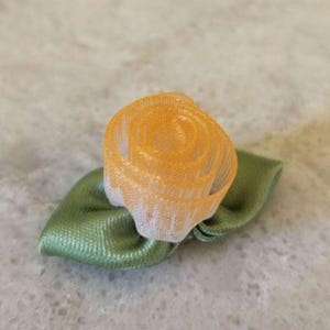May include: A small, handmade fabric flower with an orange and white rose and green leaves. The rose is made of a delicate, sheer material, and the leaves are a solid, satin-like fabric. The flower is likely used for crafts or embellishments.