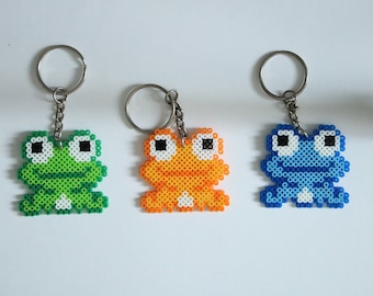 May include: Three colorful frog keychains. The keychains are made of small, colorful beads. One frog is green, one is orange, and one is blue. Each frog has a silver keyring attached.