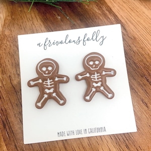 Könnte beinhalten: Ein Paar braune Skelett-Lebkuchenmann-Ohrringe mit weißen Zuckergussdetails. Die Ohrringe befinden sich auf einer weißen Karte mit dem Text "a frivolous folly" und "MADE WITH LOVE IN CALIFORNIA".