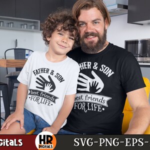 Op de afbeelding: Een wit T-shirt met de tekst "Father & Son best friends for life" en een zwart T-shirt met de tekst "Father & Son best friends for life". Beide shirts hebben een handafdruk-ontwerp.