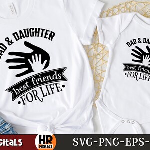 Op de afbeelding: Twee witte T-shirts met zwarte tekst en afbeeldingen. Het grotere shirt zegt "Papa & Dochter beste vrienden voor het leven" met een afbeelding van twee handen, een grote en een kleine, die elkaar aanraken. Het kleinere shirt zegt "Papa & Dochter beste vrienden voor het leven" met dezelfde handafbeelding.