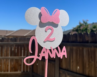 May include: A pink and white cake topper featuring a white glitter Mickey Mouse head, a pink bow, and the number 2. The name "Janna" is written in pink cursive below the number.