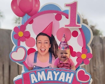 May include: A pink and blue cake topper with a photo of a girl and her mother. The topper has a pink number one, a heart, and the name "Amayah".