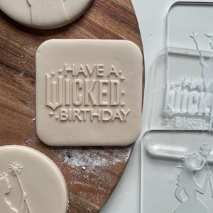 May include: A set of three cookie cutters featuring a silhouette of a woman with a wand, the text "Have a Wicked Birthday", and a silhouette of a woman with a wand. The cookie cutters are made of clear plastic and are on a wooden surface.