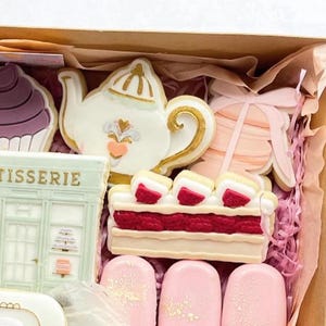 May include: A gift box filled with pink and white cookies, including a teapot cookie, a strawberry shortcake cookie, and a bow cookie. The box is lined with pink and white tissue paper.