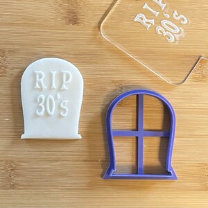 May include: A white cookie shaped like a tombstone with the text "RIP 30's" on it. A purple plastic cookie cutter shaped like a tombstone with a window design is also shown.