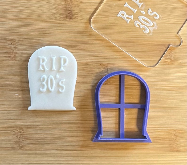 May include: A white cookie shaped like a tombstone with the text "RIP 30's" on it. A purple plastic cookie cutter shaped like a tombstone with a window design is also shown.