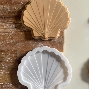 May include: Two seashell-shaped cookie cutters. One is a light tan colour, and the other is white. The cutters are on a wooden surface. The seashell design features radiating lines.