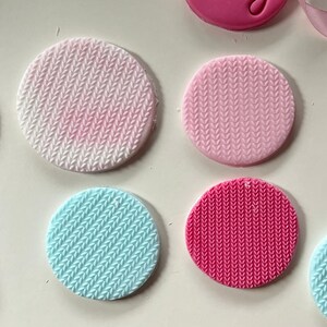 May include: Four round fondant cake toppers with a knitted pattern. The toppers are in pink, light blue, and a darker pink.