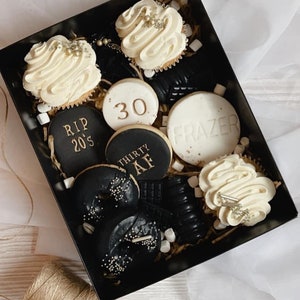 May include: A black gift box filled with a variety of black and white cookies and cupcakes. The cookies have gold lettering that says "RIP 20's", "30", "THIRTY AF", and "FRAZER". The cupcakes have white frosting with gold sprinkles.