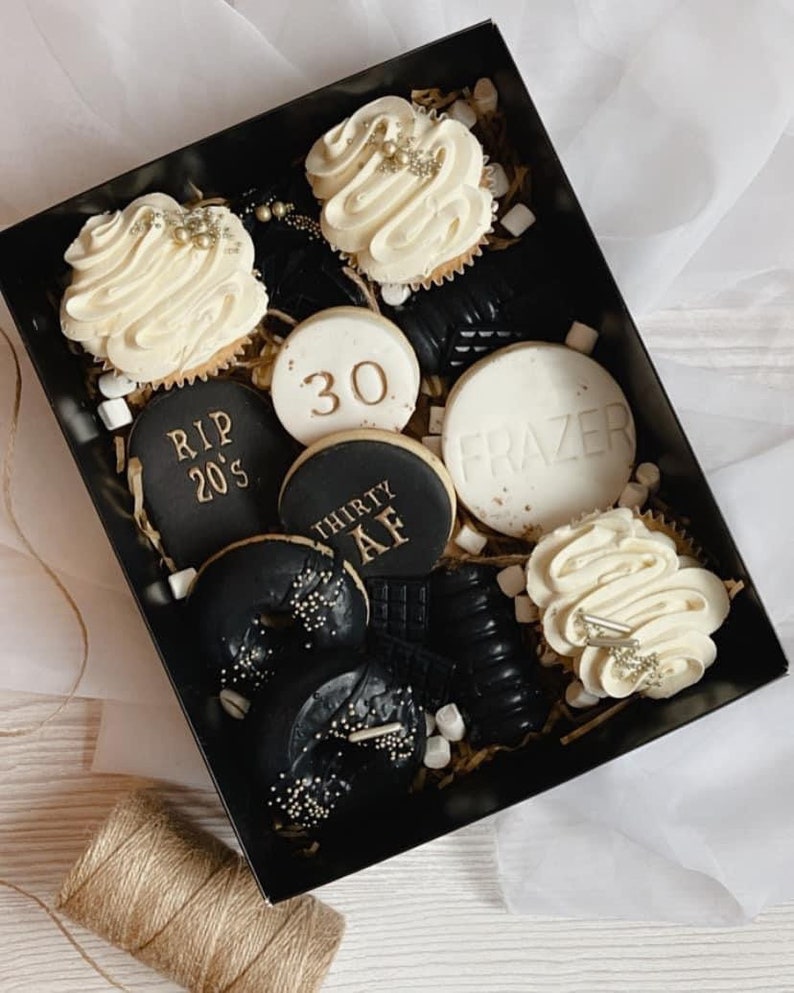 May include: A black gift box filled with a variety of black and white cookies and cupcakes. The cookies have gold lettering that says "RIP 20's", "30", "THIRTY AF", and "FRAZER". The cupcakes have white frosting with gold sprinkles.