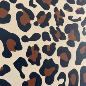May include: Close-up of a cream-coloured surface with a repeating leopard print pattern. The pattern features dark navy blue and brown spots of varying sizes and shapes, creating a bold and stylish design.