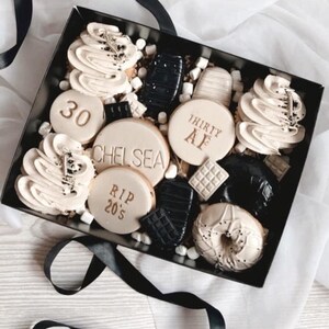 May include: A black gift box filled with a variety of cookies and treats. The cookies are decorated with white frosting and gold lettering. Some of the cookies have the words "30", "Chelsea", "RIP 20's", and "Thirty AF" written on them.