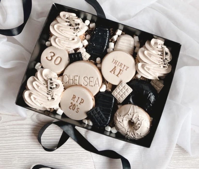 May include: A black gift box filled with a variety of cookies and treats. The cookies are decorated with white frosting and gold lettering. Some of the cookies have the words "30", "Chelsea", "RIP 20's", and "Thirty AF" written on them.