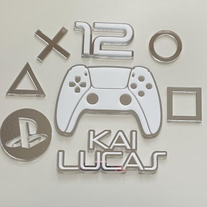 May include: A set of silver and white gaming-themed decorations, featuring a controller, symbols, the number 12, and the name "KAI LUCAS". The items are displayed on a light background, ideal for gamers.
