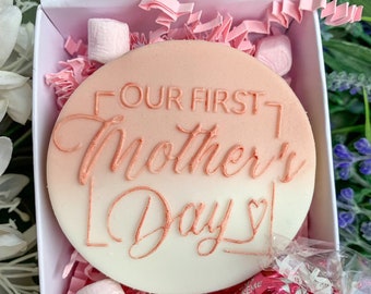 May include: A white and pink round cake decorated with the words "Our First Mother's Day" in a rose gold color. The cake is in a white gift box with pink tissue paper and other treats, including pink and white marshmallows, a red candy, and a pink chocolate.