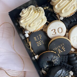 May include: A black gift box filled with a variety of treats, including cupcakes, cookies, and donuts. The cupcakes are topped with white frosting and gold sprinkles. The cookies are decorated with gold lettering that says "RIP 20's" and "Thirty AF". The donuts are black with gold sprinkles.