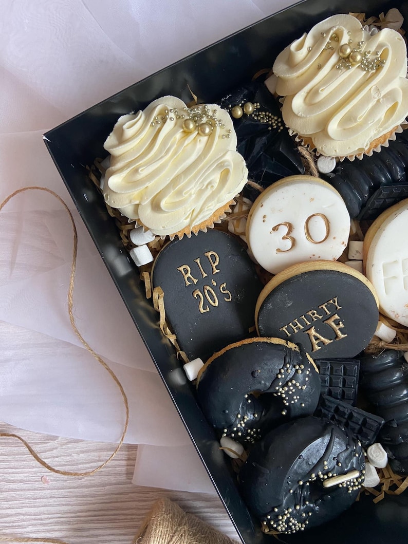 May include: A black gift box filled with a variety of treats, including cupcakes, cookies, and donuts. The cupcakes are topped with white frosting and gold sprinkles. The cookies are decorated with gold lettering that says "RIP 20's" and "Thirty AF". The donuts are black with gold sprinkles.