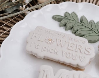 May include: A white ceramic plate with three decorative cookies. One cookie is a rectangle with the text "If mums were flowers I'd pick you" and a small flower design. Another cookie is a leaf design. The third cookie is a letter "M" with a flower design.